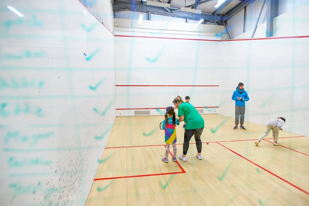 Junior squash session on court with coach giving one-on-one support, viewed through a glass wall with a checklist.