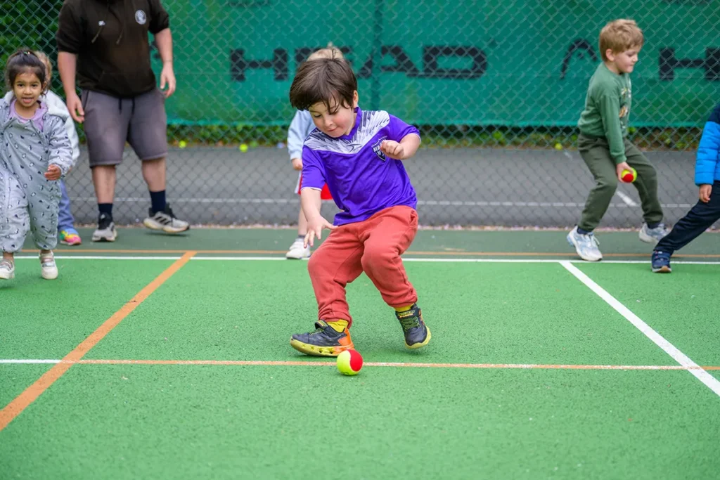 Young children playing a coordination ball game on court during a Coolhurst junior tennis session, with coaches in the background.