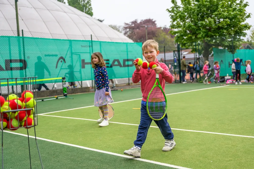 Smiling young boy holding soft tennis balls and a racket during a kids' session at Coolhurst, with other children playing in the background.