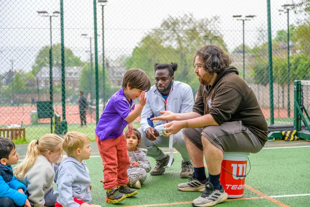 Young children sitting on a tennis court listening to two coaches, one handing out stickers or rewards, during a kids' tennis session.