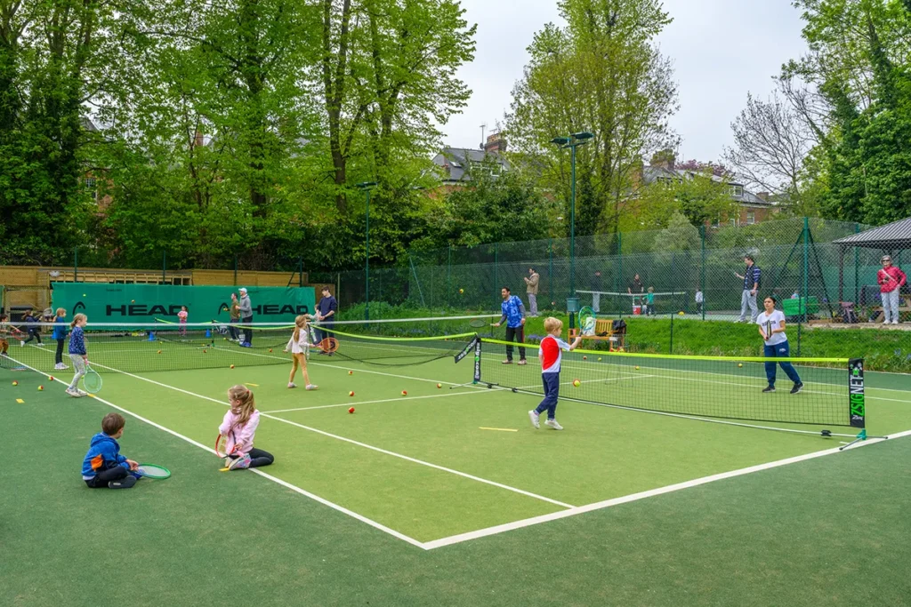 Young children enjoying a group tennis coaching session on outdoor mini courts, surrounded by trees and watching adults.