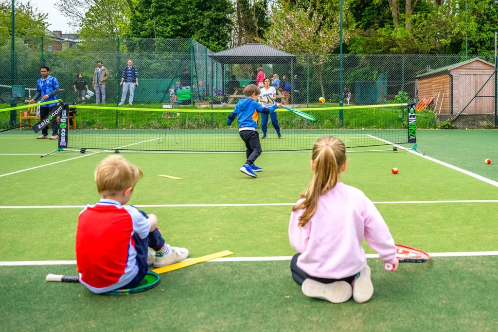 Children taking part in a mini tennis session on a green outdoor court, with coaches and parents watching in the background.