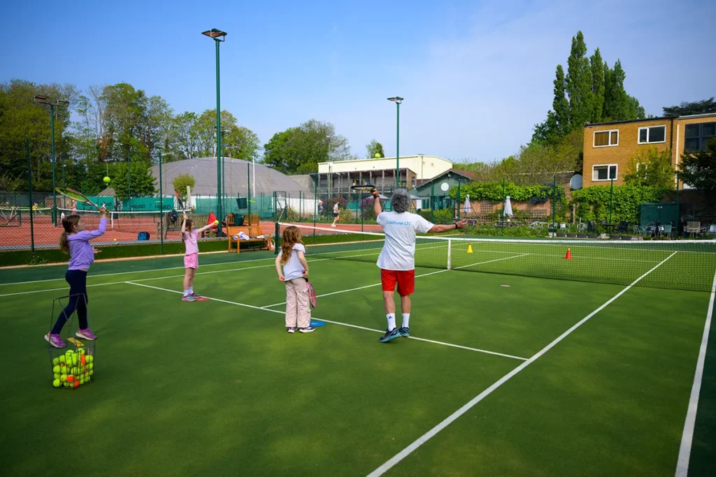 Junior tennis coaching session on a green court with three young girls and a coach practicing forehands.