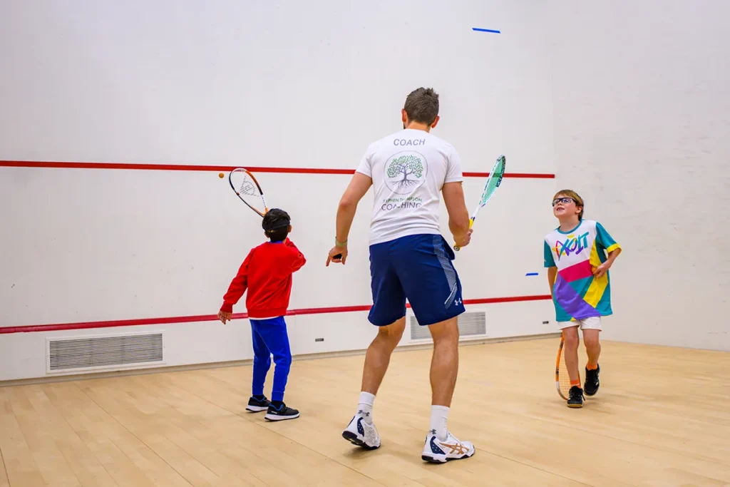 Squash coach working with two young children on court, one preparing to hit the ball.