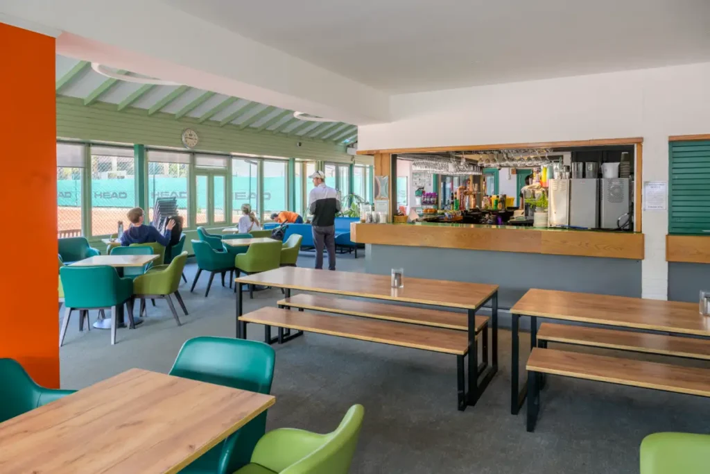 Spacious clubhouse café with a mix of bench and armchair seating, bar counter, and view of outdoor tennis courts through large windows.