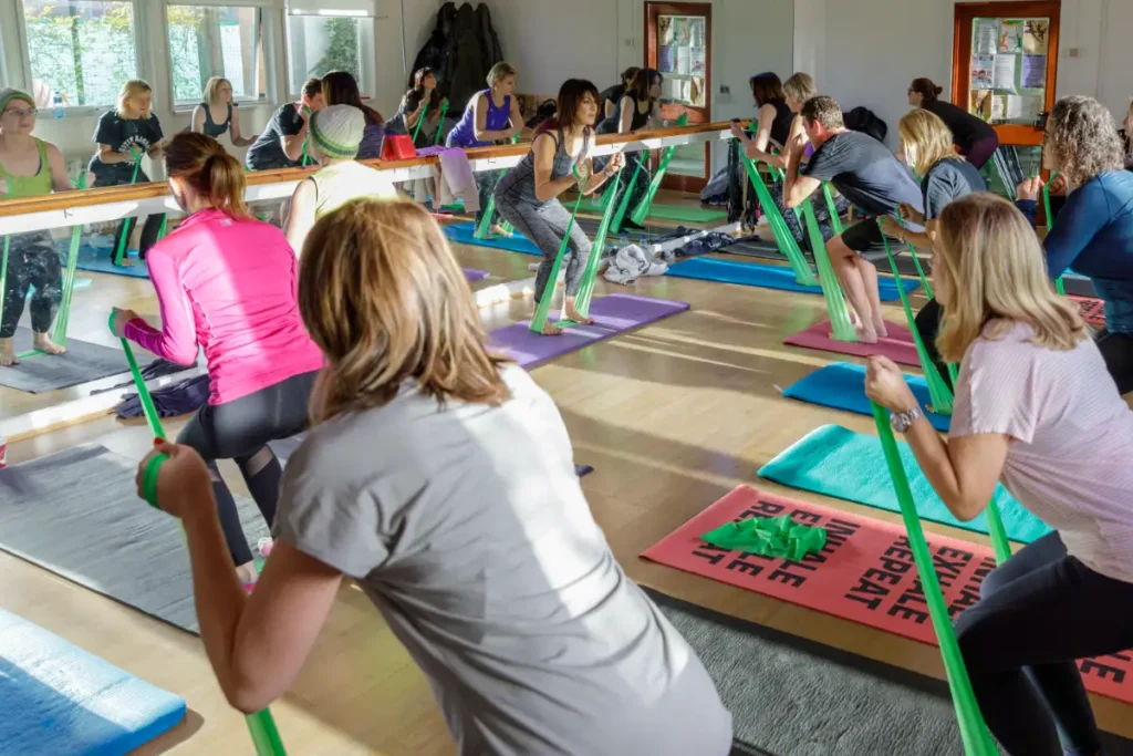 Large group fitness class using green resistance bands in front of mirrors on yoga mats.