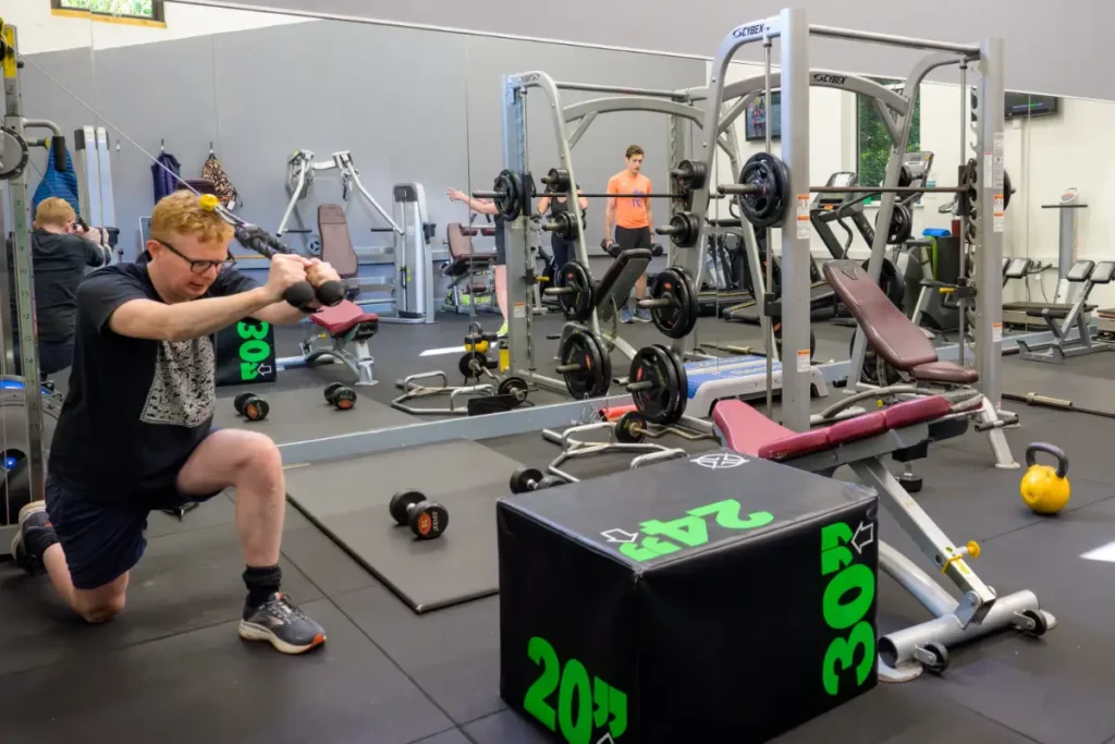 Person kneeling while using a cable machine in a modern fitness studio, surrounded by various workout equipment and mirrored walls.