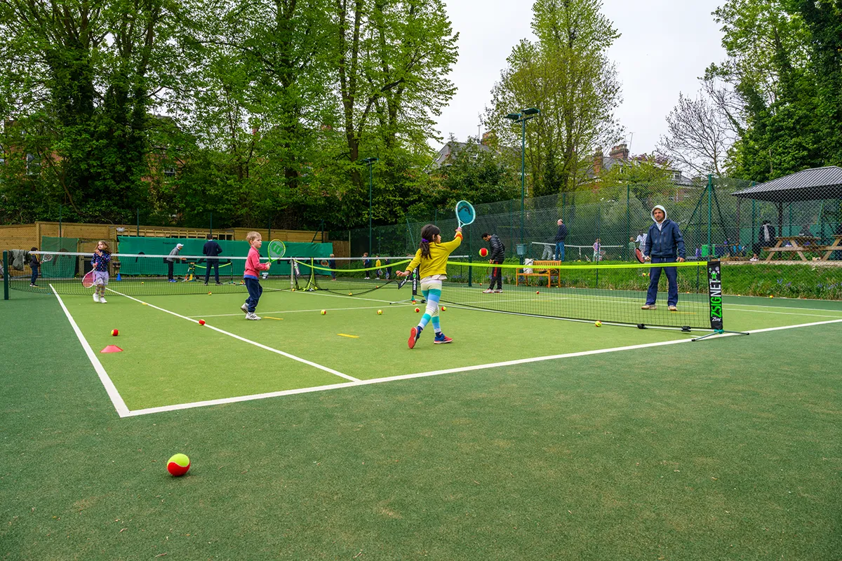 Children playing tennis on an outdoor mini court with a low net. A coach stands on the opposite side, watching and instructing the kids. Trees and fencing surround the court.