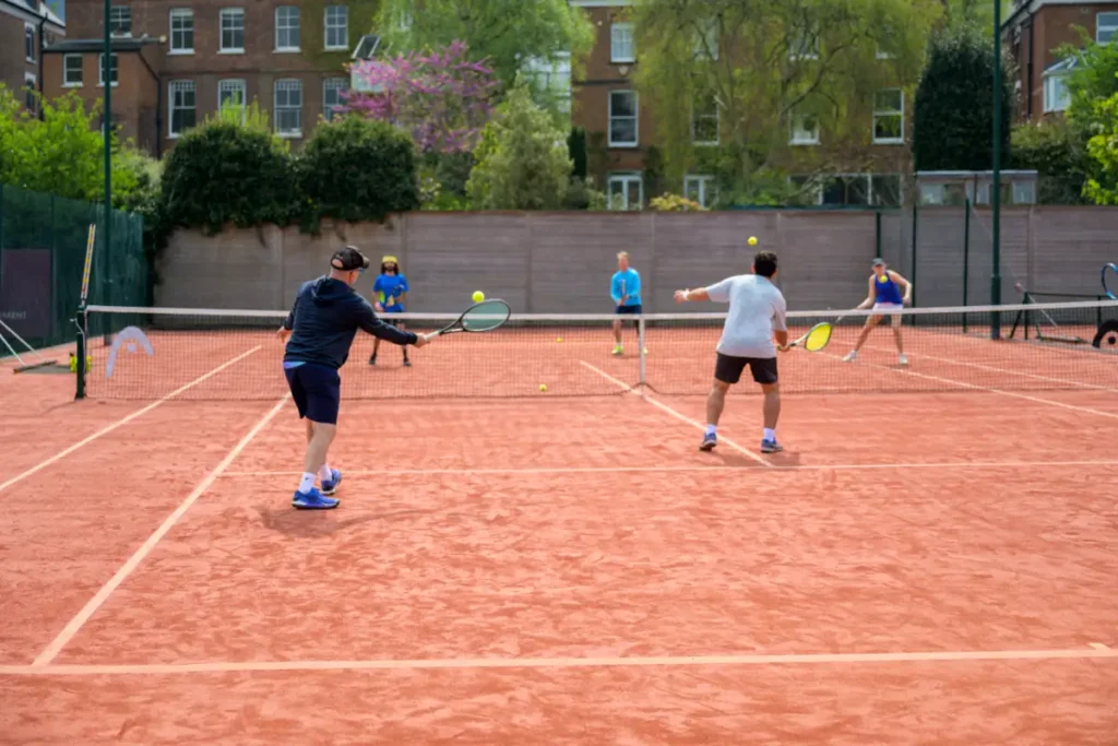 Players practicing doubles on a red clay court, with several tennis balls mid-air and others watching in the background.