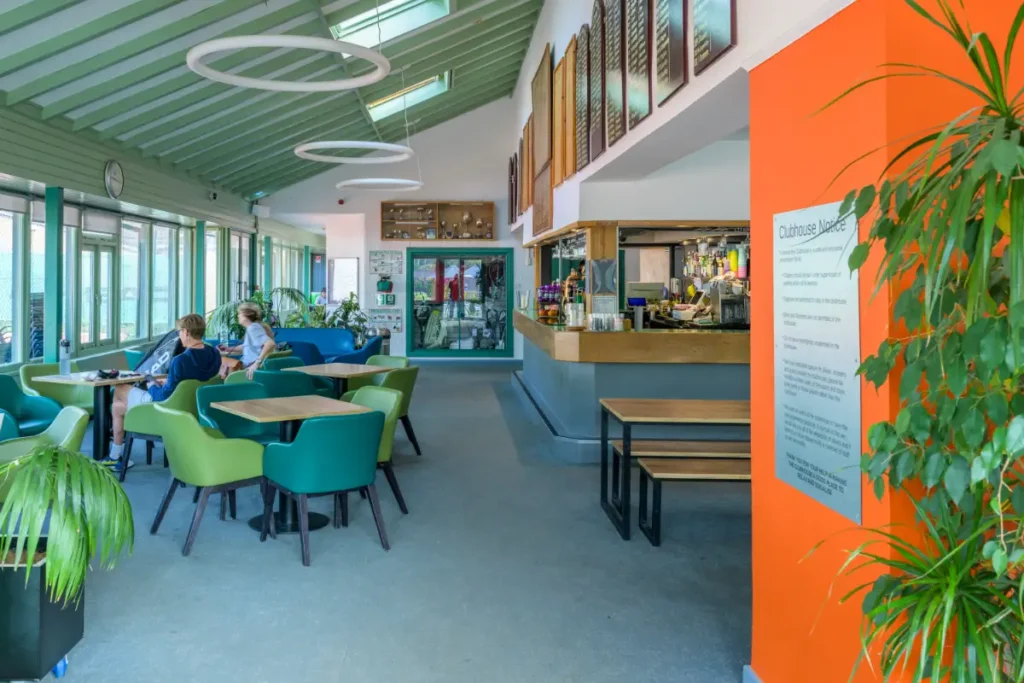 Modern clubhouse interior with colourful chairs, bar area, and large windows overlooking tennis courts.