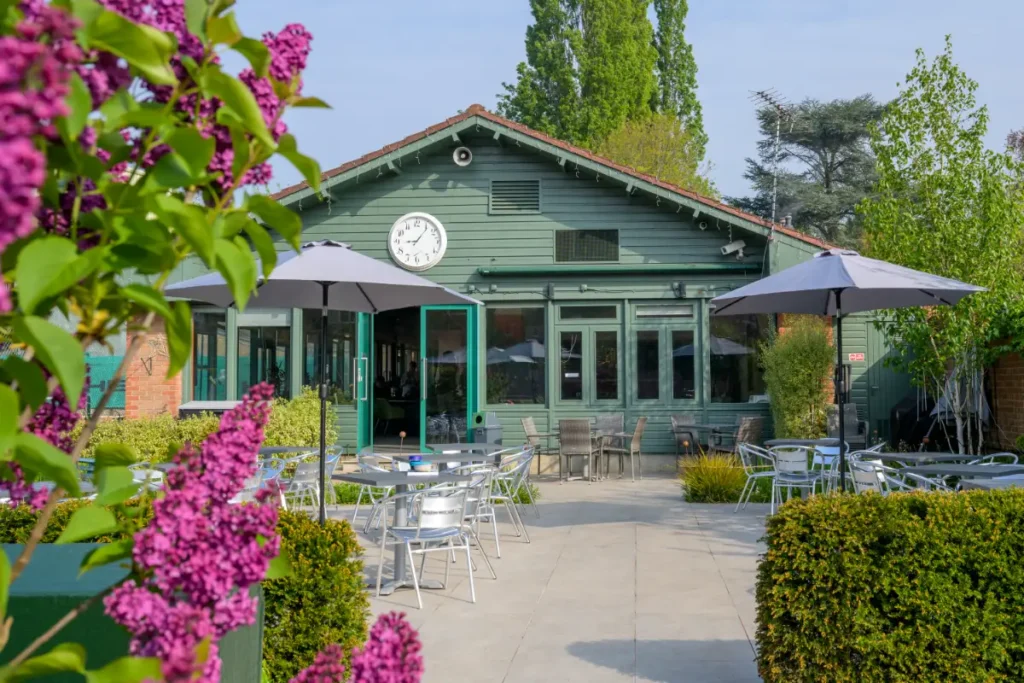 Green-painted clubhouse with terrace seating, umbrellas, and blooming purple flowers in the foreground.