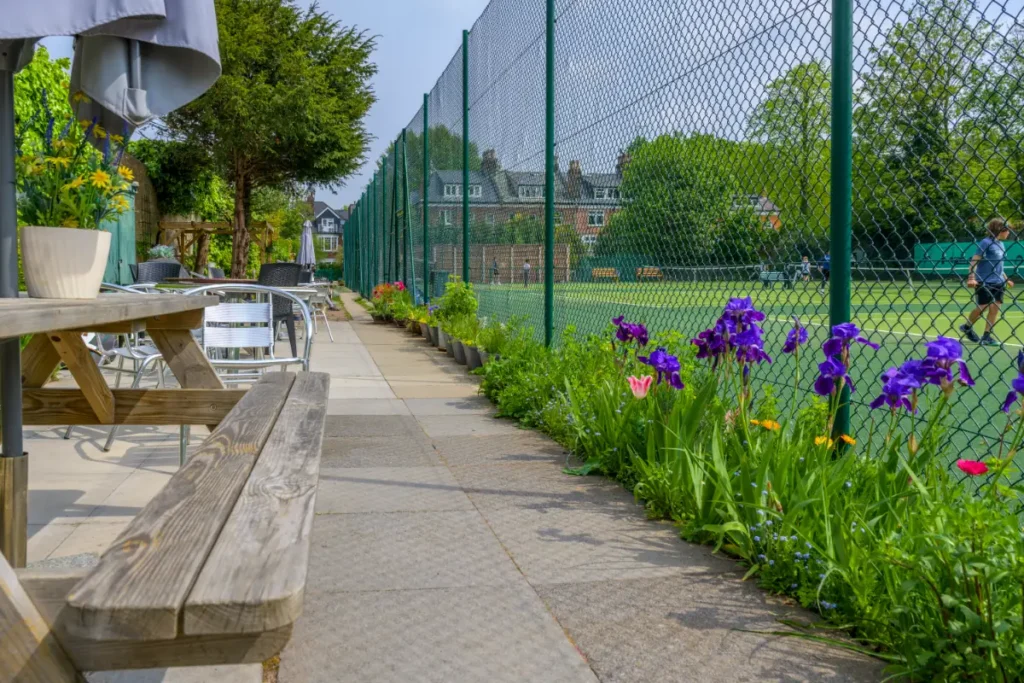 Courtside seating area with picnic benches and metal chairs alongside a flower-lined tennis court fence.