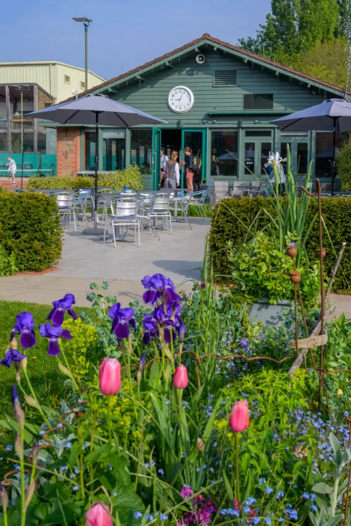 Clubhouse with a clock above the entrance, outdoor seating, and a foreground garden of tulips and irises.