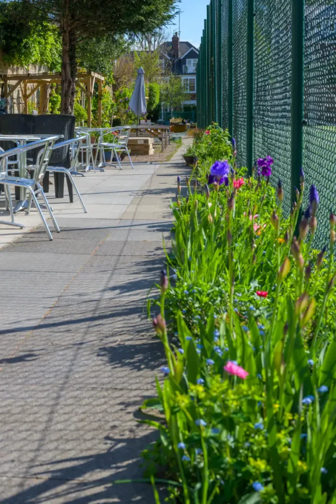 Outdoor seating area with metal chairs and tables alongside a flower bed and tennis court fencing on a sunny day.
