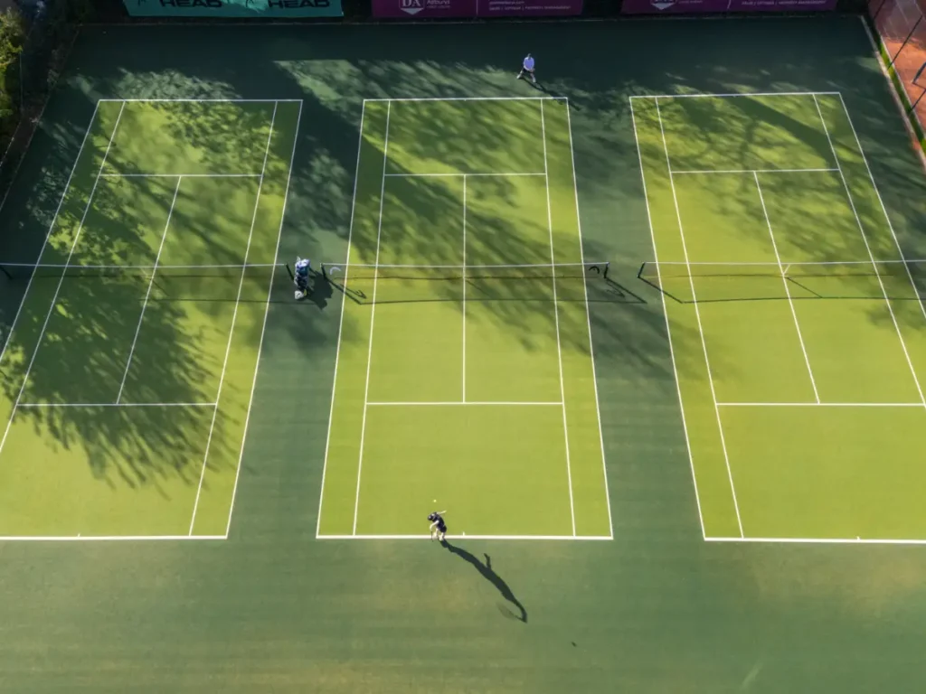 Overhead view of three green artificial grass tennis courts with players on court and shadows cast by nearby trees.