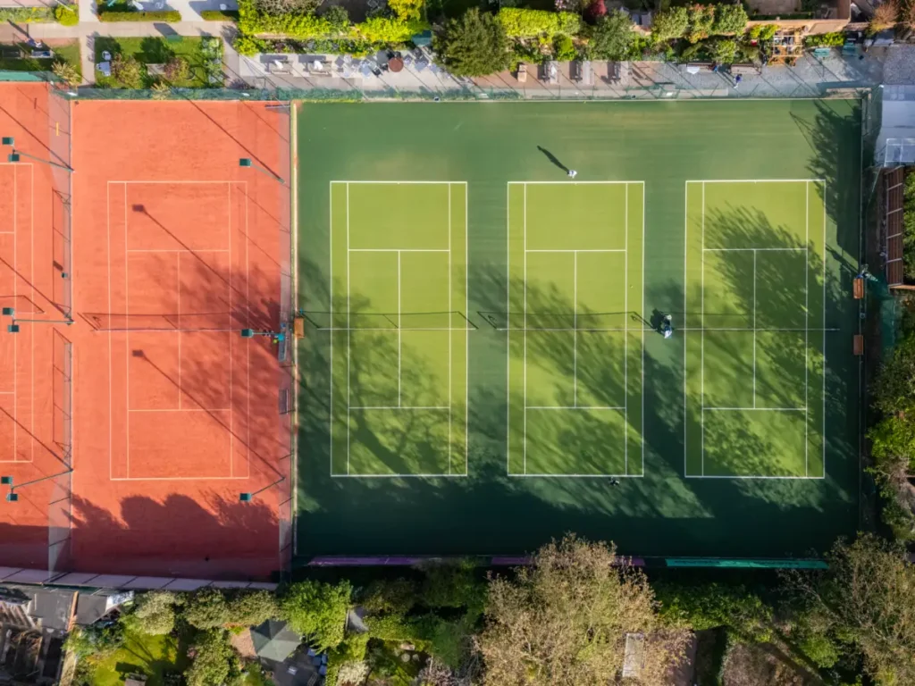 Aerial view of adjacent red clay and green artificial grass tennis courts surrounded by trees and seating areas.
