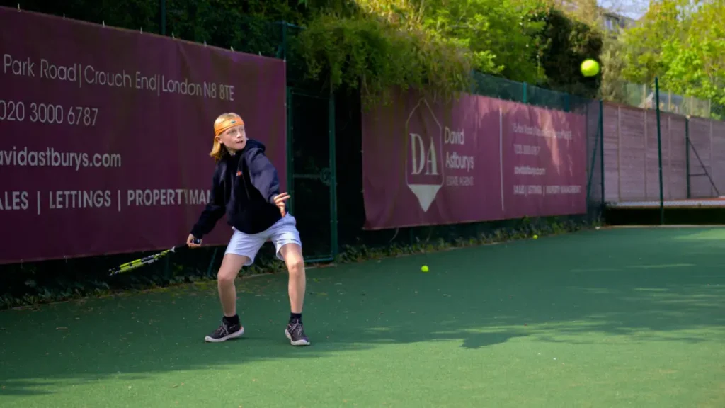 Young tennis player in a hoodie and headband preparing to return a ball on an outdoor green court.