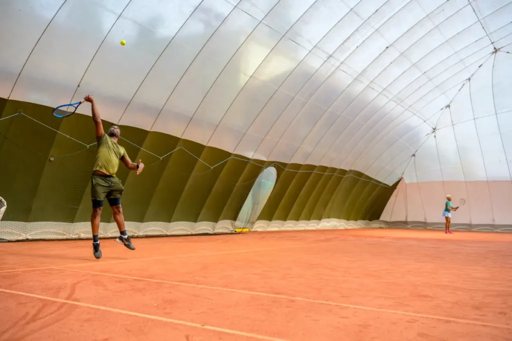 Man in green shirt serves the ball on a full-size indoor clay court under a white inflatable dome, while his opponent waits across the net.