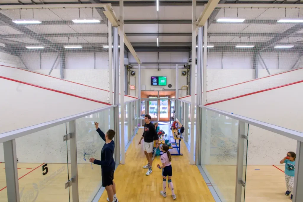 Junior squash players and a coach in a corridor between glass-backed squash courts at an indoor club.
