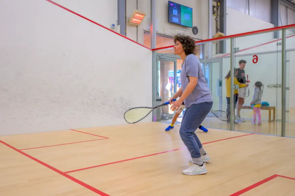 Woman prepares to return a squash shot on a well-lit court, with families and children visible through the glass wall behind her.