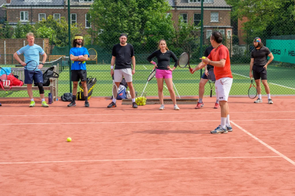 Coach in red shirt explaining a tennis drill to a group of players on a red clay court, with rackets and baskets of balls nearby.