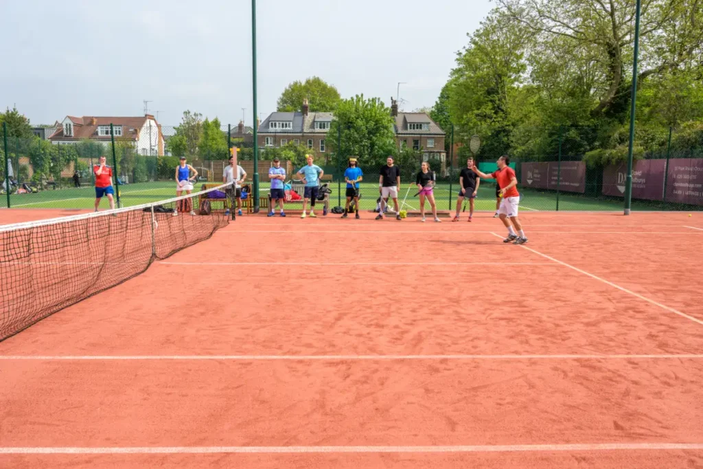 Group of players standing on a red clay tennis court watching a coach demonstrate a shot during a session.