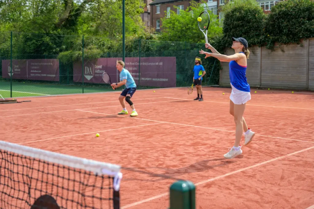 Three players on a red clay tennis court during a coaching session, with one preparing to serve.