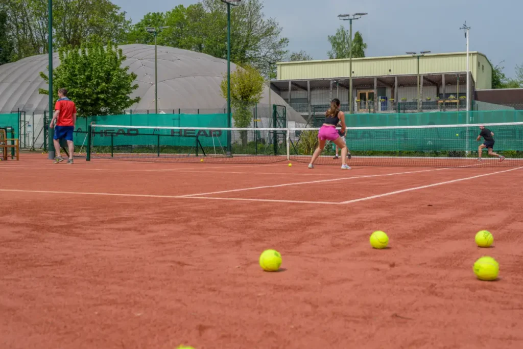 Tennis players rallying on a red clay court with scattered tennis balls in the foreground and practice domes in the background.