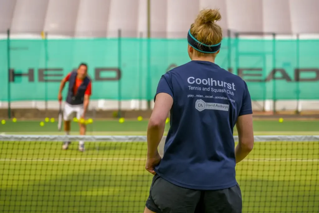 Person wearing a Coolhurst Tennis and Squash Club shirt on court, facing a coach with tennis balls scattered on the surface.