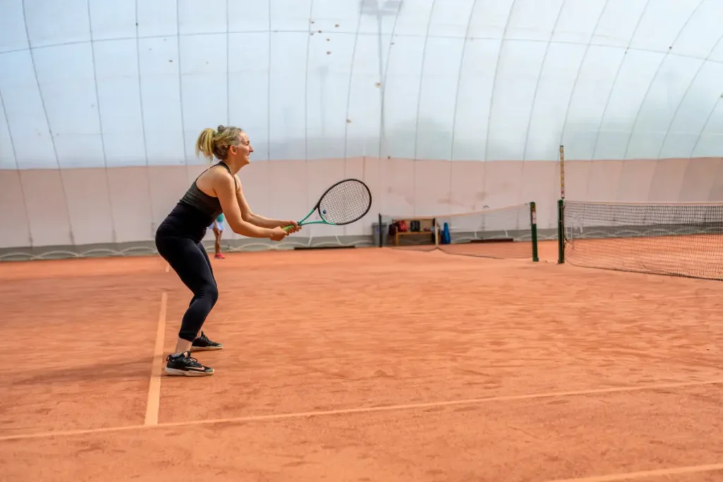 Female tennis player in black outfit on a red clay court inside an inflatable dome, preparing to hit the ball with a two-handed grip.