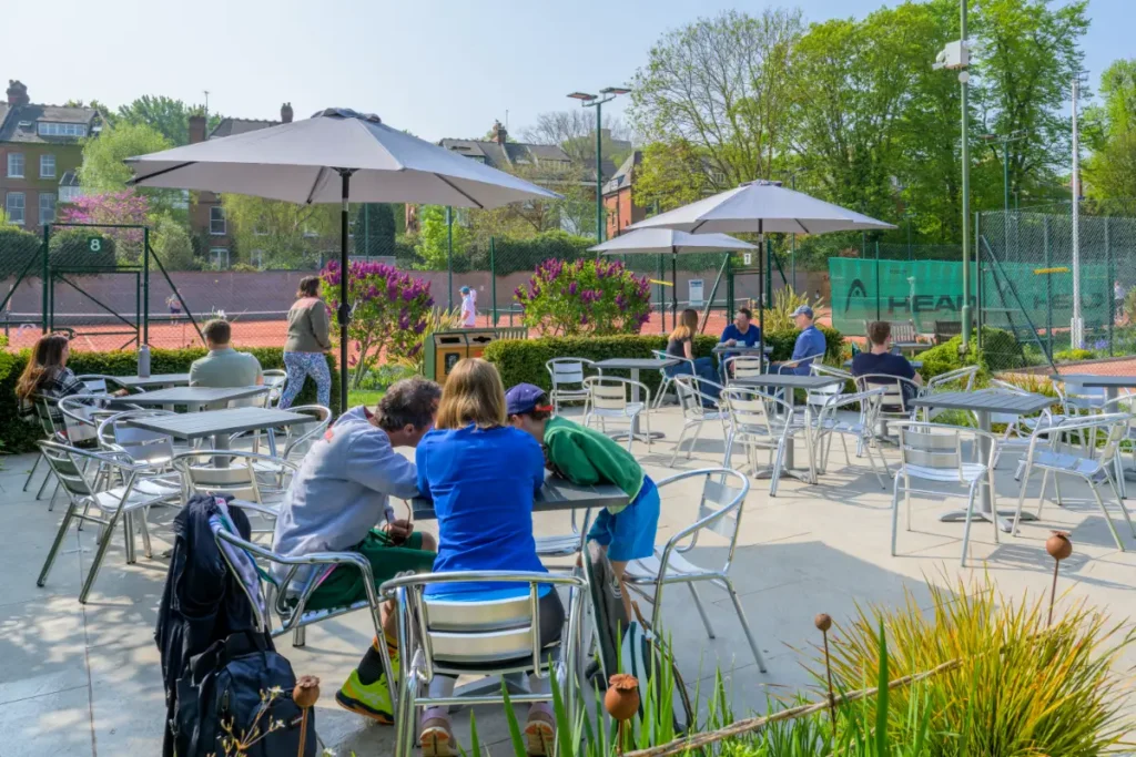 Outdoor seating area with parasols and metal chairs, overlooking tennis courts in use, surrounded by greenery and flowers.