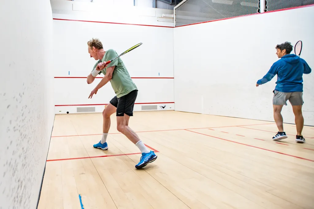 Two men playing squash in an indoor court, one in a green shirt preparing to hit the ball near the front wall, the other in blue watching closely.