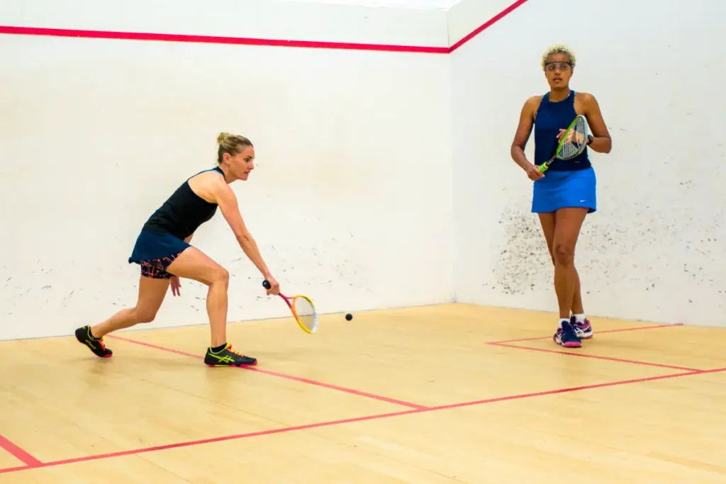 Two women playing squash, one reaching to hit the ball with a forehand while the other watches.