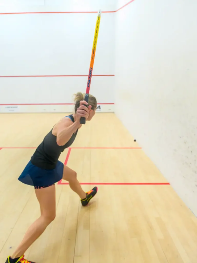 Female squash player in mid-swing, stretching toward the ball inside a white-walled court.