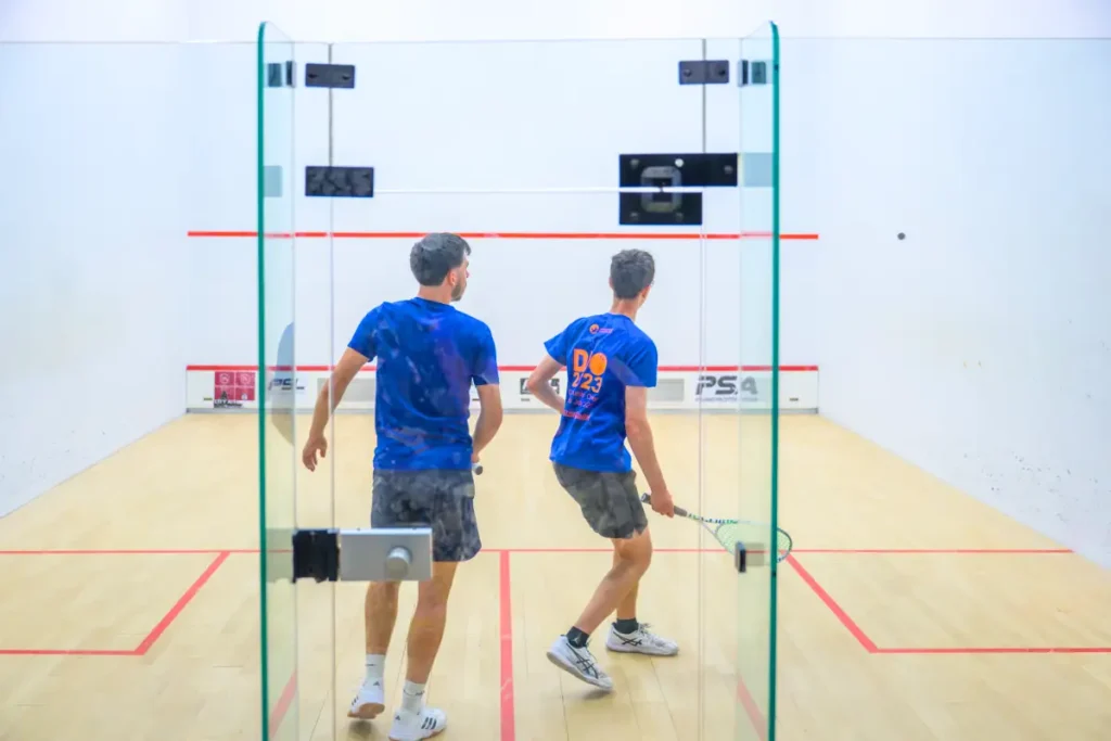 Two players in blue shirts competing on a glass-back squash court, mid-rally.