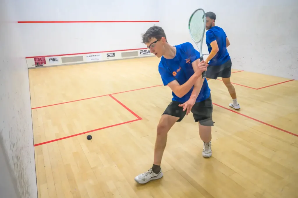 Two young men in blue shirts playing squash on an indoor court, one preparing to return the ball.