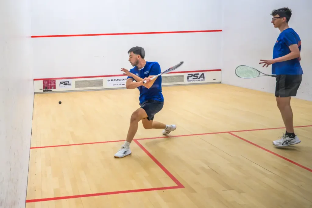 Squash player in mid-swing returning a low ball while another player looks on.