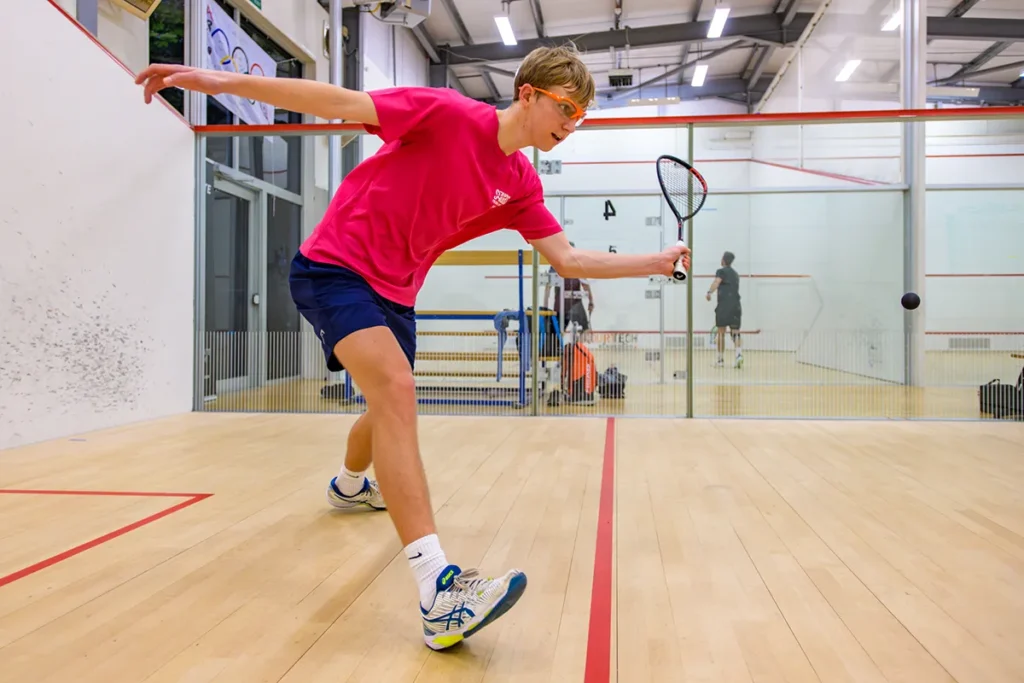 Young man wearing a bright pink shirt and protective glasses stretches to hit the squash ball inside a glass-walled indoor court.