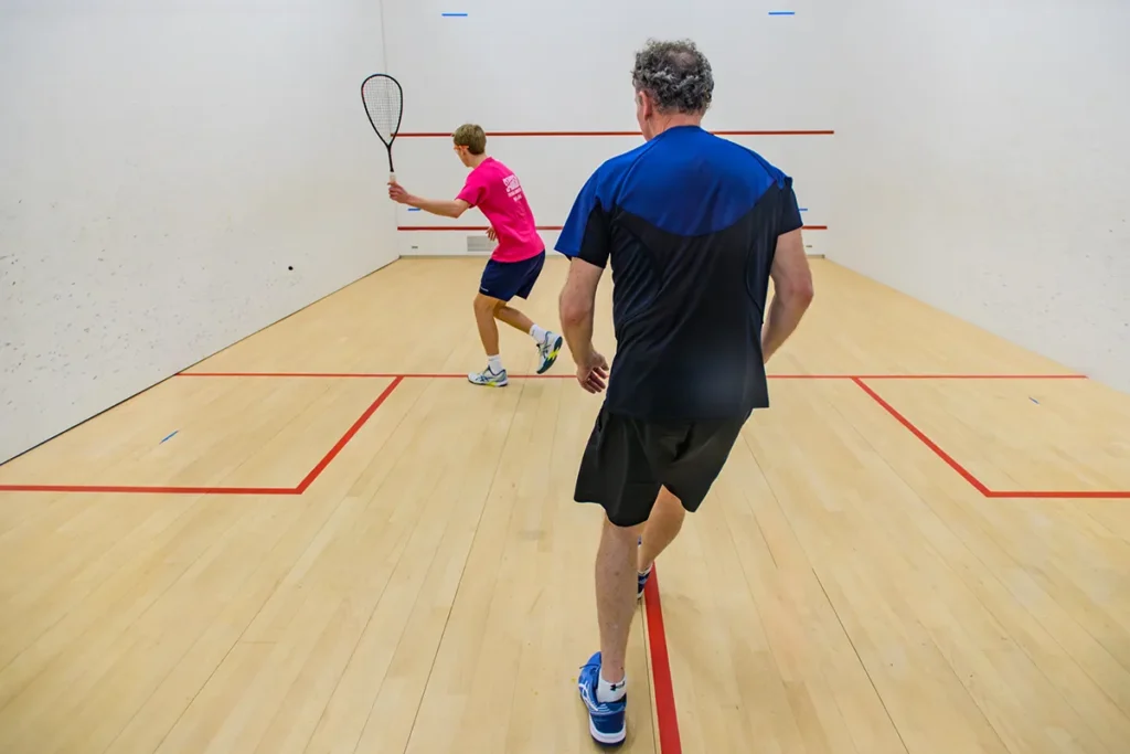 Two men playing squash in an indoor court, one preparing to hit the ball while the other follows.