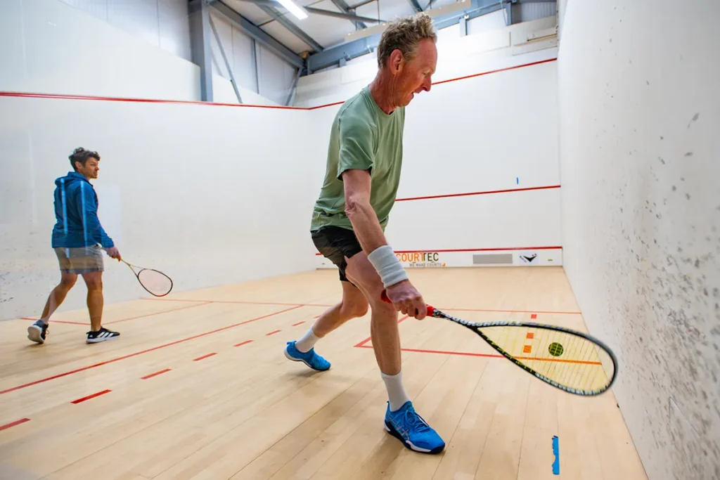 Two men playing squash in an indoor court, one preparing to hit the ball close to the wall.