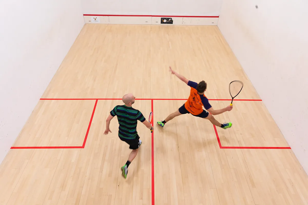 Overhead view of two men playing squash on an indoor court, one lunging to hit the ball.