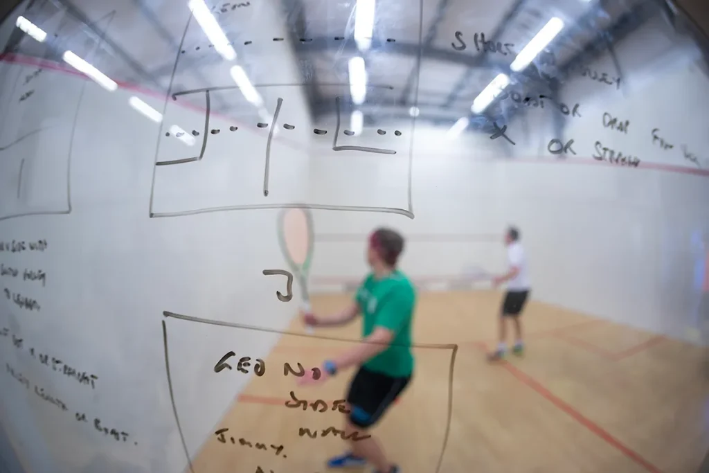 Squash court with tactical notes written on the glass wall, players warming up or practicing in the background.
