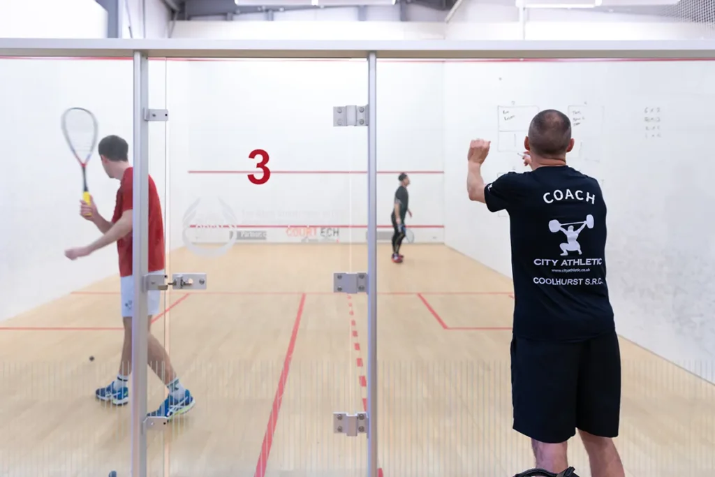 Squash court view through glass with a coach drawing tactics on the wall and players practicing in the background.