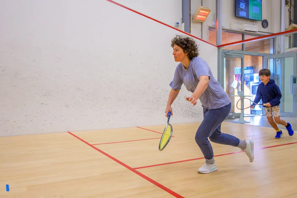 Woman and child playing squash on a glass-backed indoor court, mid-rally.