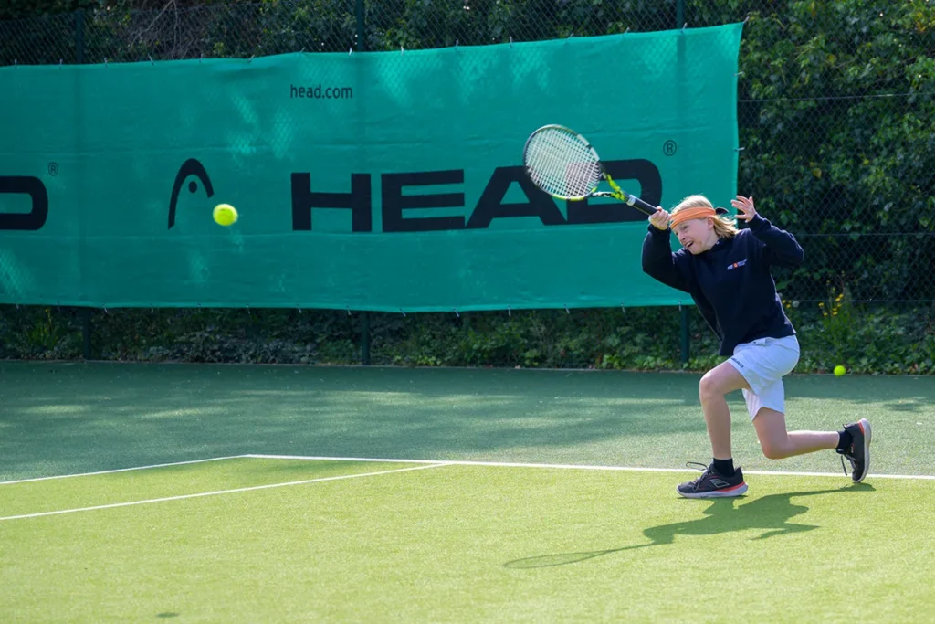 Young tennis player lunging to hit a forehand on green outdoor court at Coolhurst Tennis Club with windscreen in background.