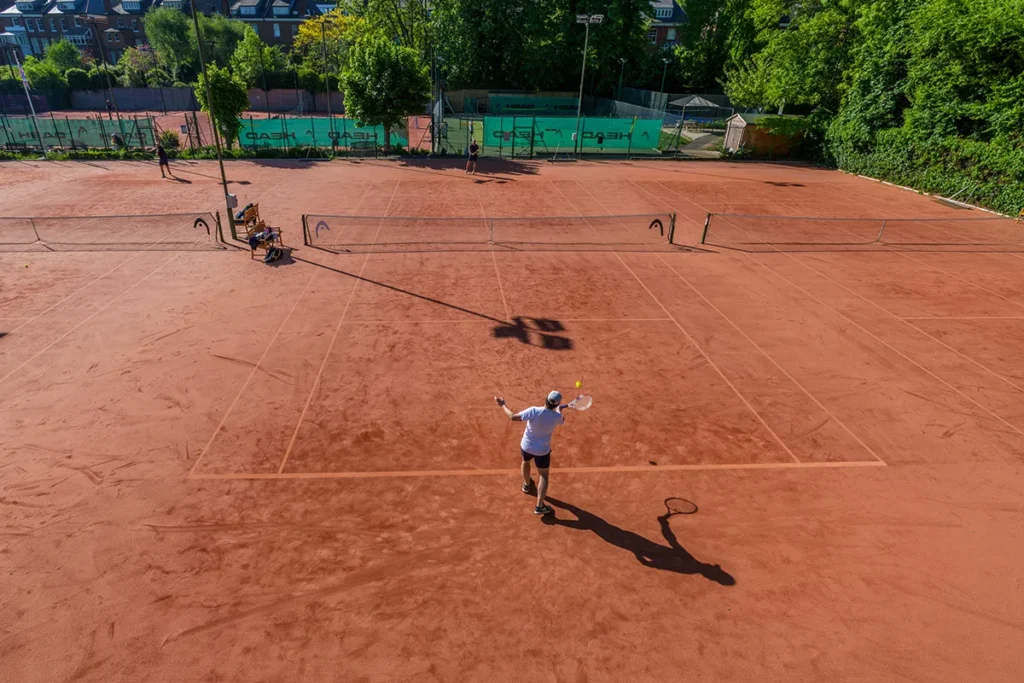 Aerial view of players engaged in a tennis match on a clay court at Coolhurst Tennis Club, surrounded by trees and fencing.