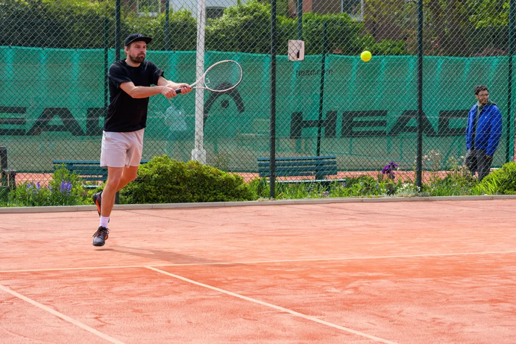 Male tennis player in black shirt and white shorts hitting forehand on red clay court at Coolhurst Tennis Club with fencing in the background.