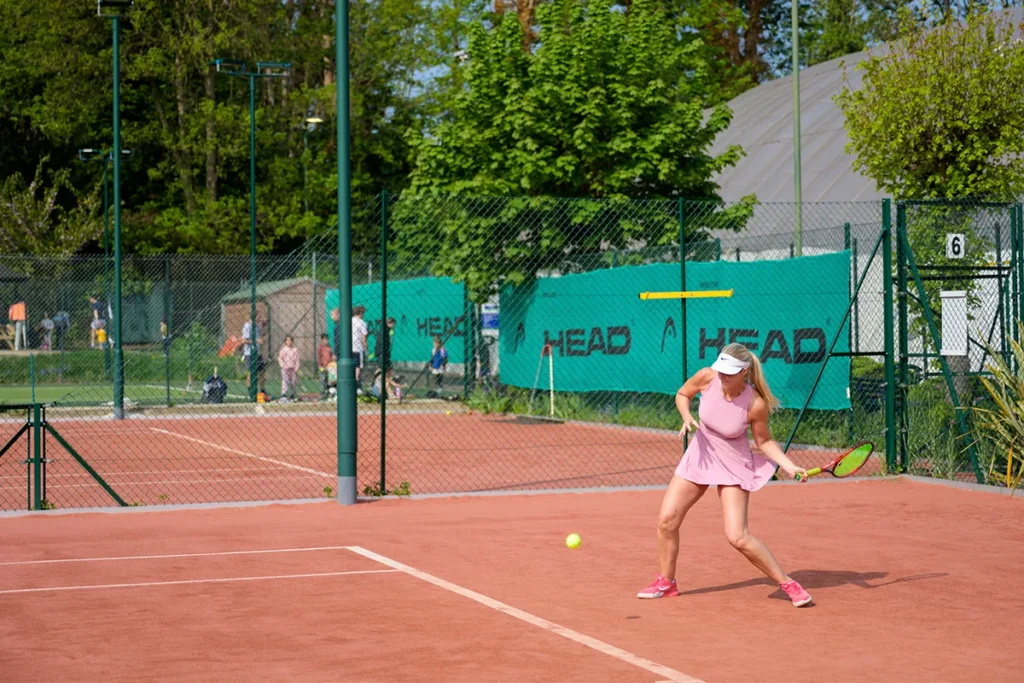 Female tennis player in pink dress practicing forehand shot on red clay court at Coolhurst Tennis Club with outdoor courts and fencing in the background.