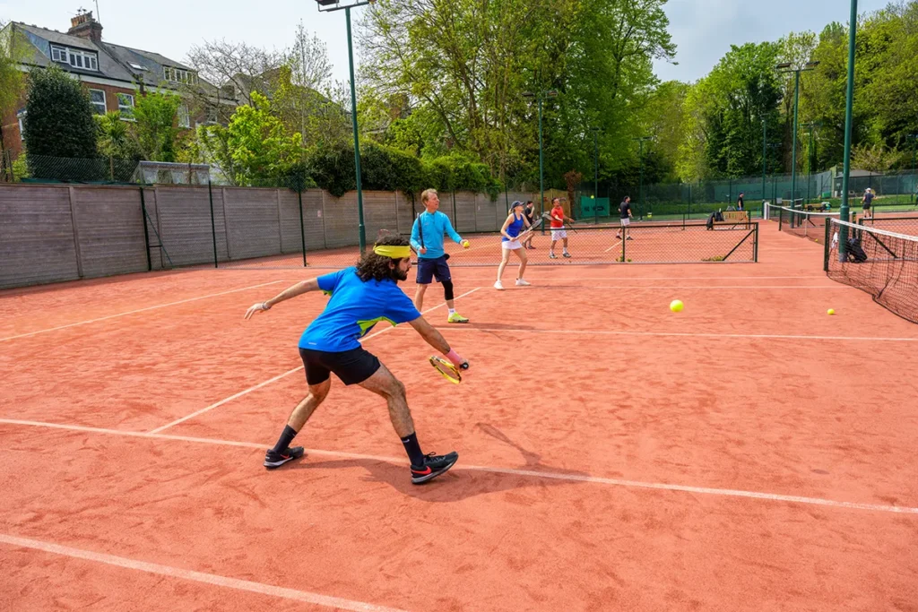 Group of tennis players on a red clay court at Coolhurst Tennis Club. One player in blue is lunging forward to hit the ball, while others prepare for the next shot on adjacent courts. Trees and fencing surround the area.