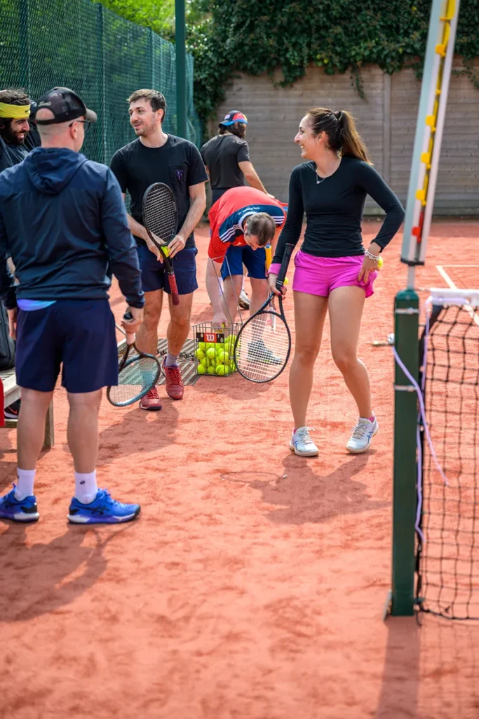 A group of tennis players gathers on a red clay court at Coolhurst Tennis Club, chatting and preparing for practice. One woman in black top and pink shorts smiles, while others hold rackets near a basket of tennis balls.